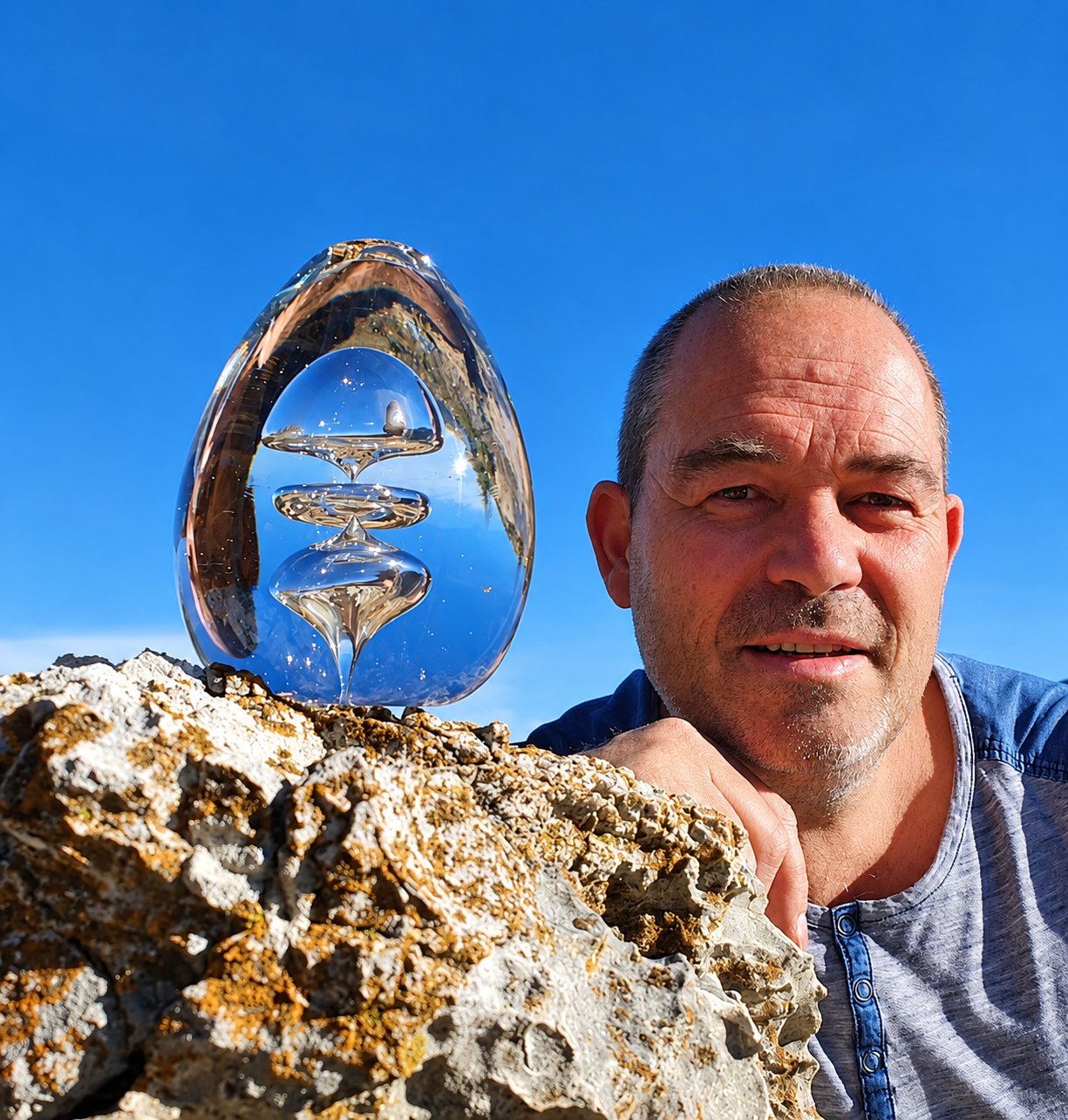 Un homme au crâne rasé sourit près d'une sculpture en verre transparent en forme d'œuf posée sur un rocher, sous un ciel bleu.