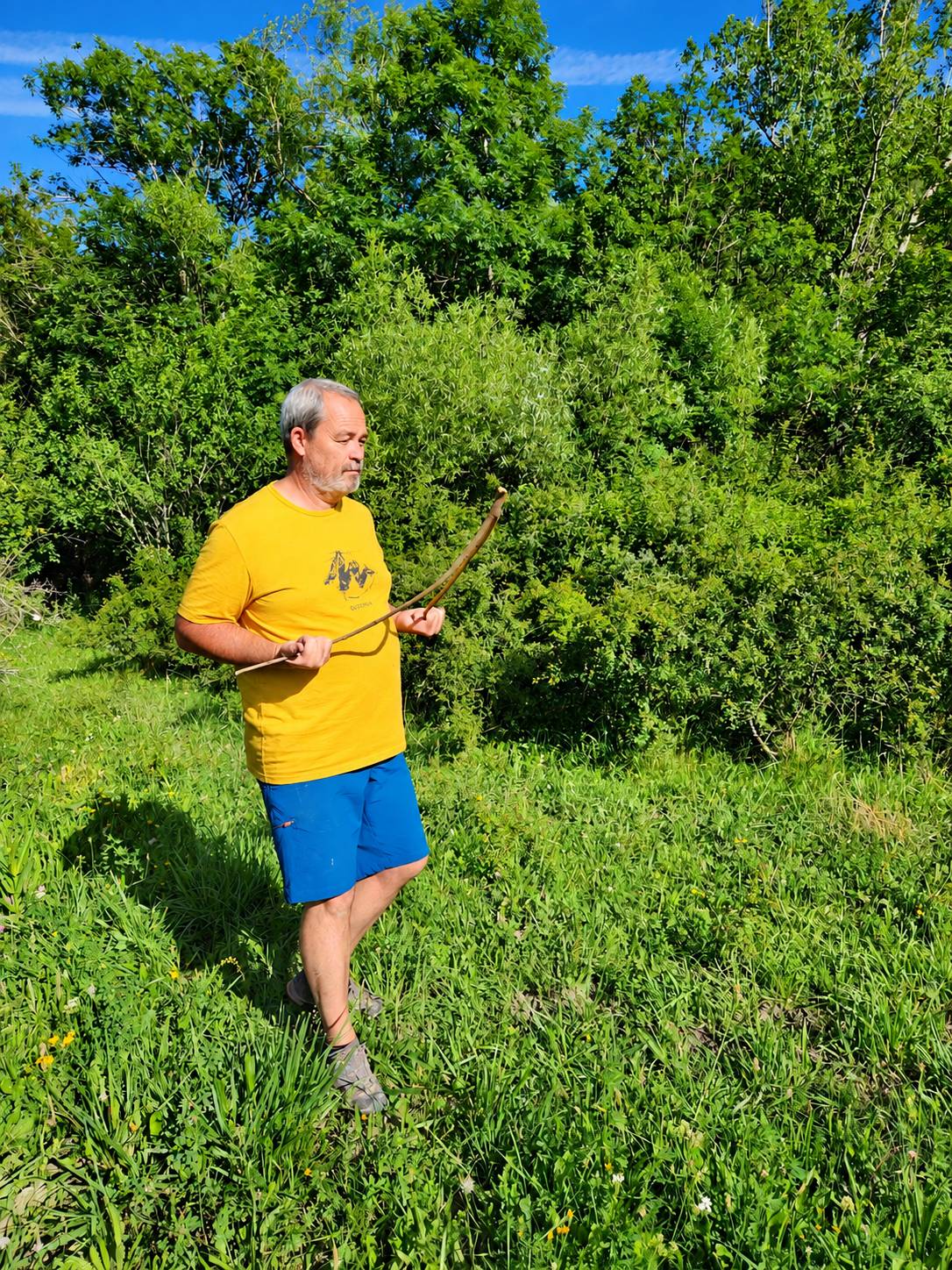 Un homme aux cheveux gris, vêtu d’un t-shirt jaune et d’un short bleu, tient un arc en bois courbé dans une prairie entourée d’arbres.